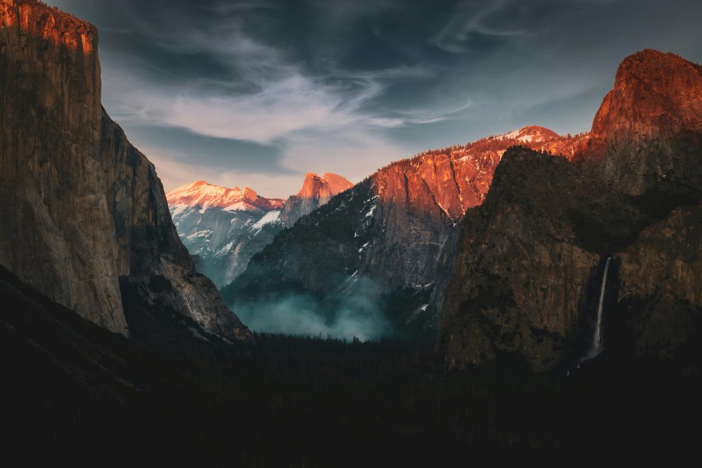 A sunset view of Yosemite National Park in the United States. The sun is going down, and there's a moody dark sky. Most of the mountain range is in shadow, and you can see mist coming off the forest at the bottom of the image.