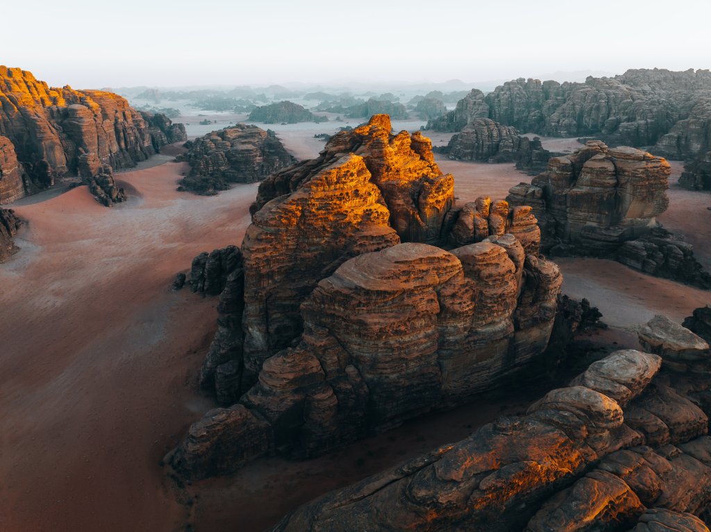 Sandstone plateau in the Hisma Desert, Saudi Arabia. It's early morning, and the sun is casting a golden hue on the top of the stone structures. It's misty in the far distance.