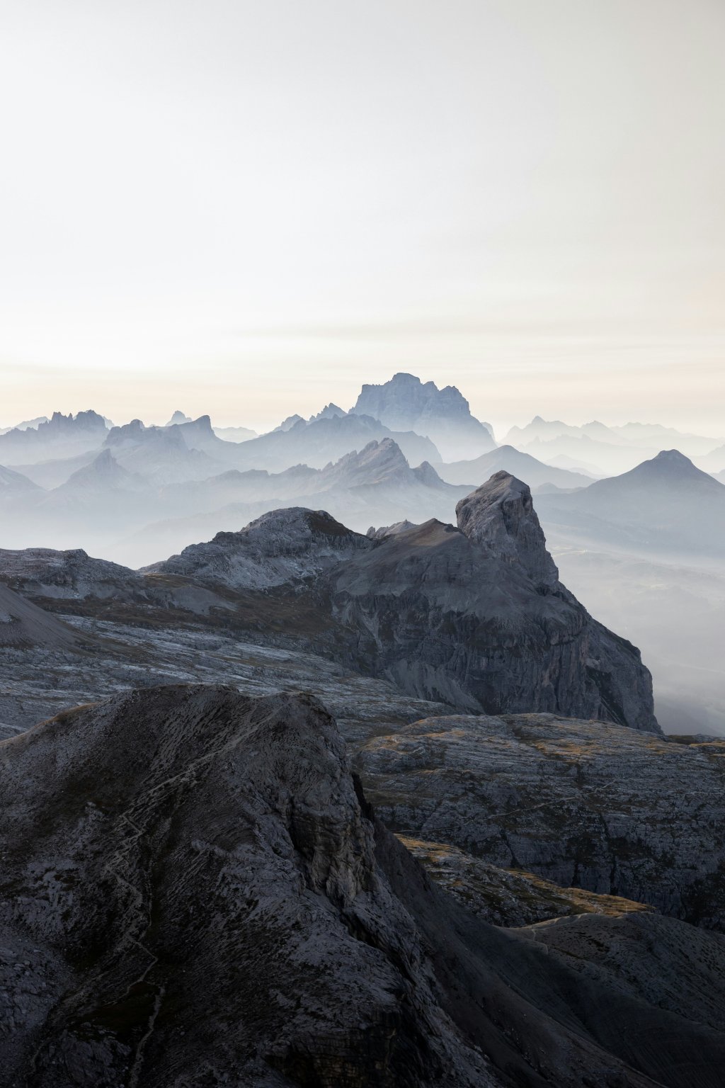 A snow-covered mountain under a cloudy sky during daytime in Puezgruppe. The lighting is bleak and there are other mountains in the background covered in mist.