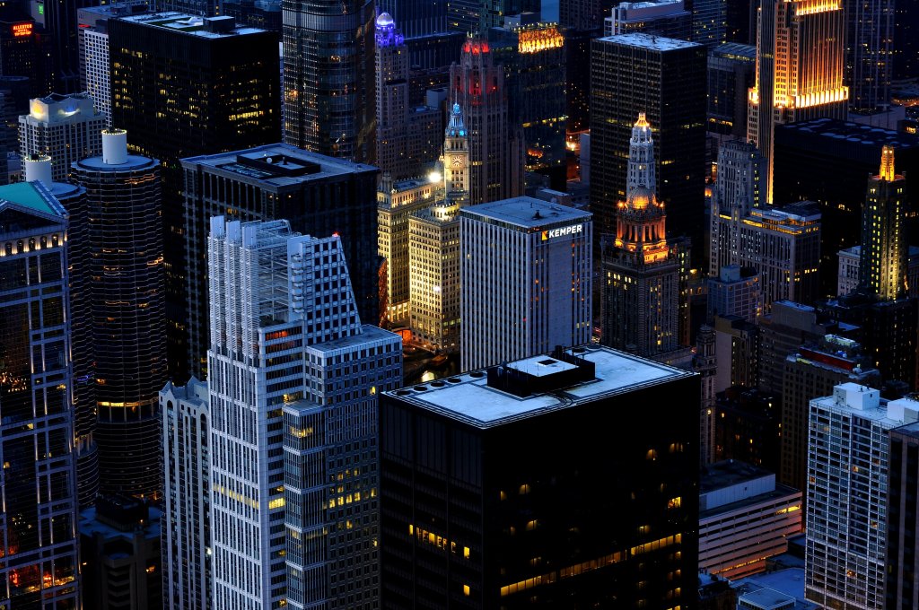 An aerial view of Chicago skyscrapers in the early evening. Various yellow uplights are shown across the buildings, which look quite pretty, and there's a blue hue.