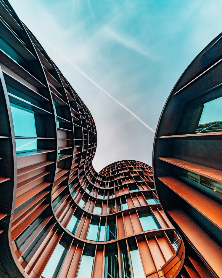 The Axel Towers in Copenhagen, Denmark. The building is distinctively wavey, with a grid-like structure and tall slits for windows. The building is a rusty colour and the camera is facing up to the light blue sky.