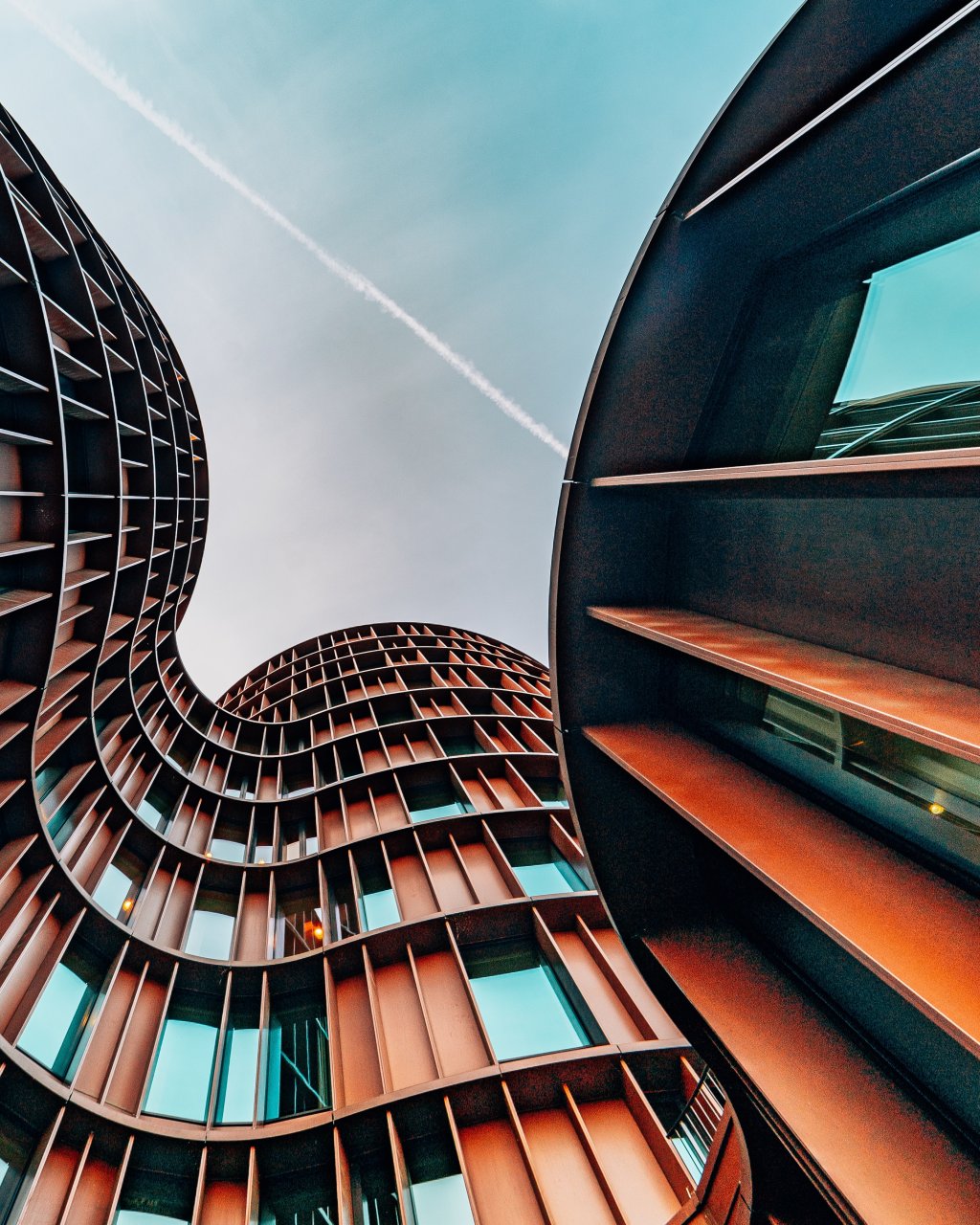 The Axel Towers in Copenhagen, Denmark. The building is distinctively wavey, with a grid-like structure and tall slits for windows. The building is a rusty colour and the camera is facing up to the light blue sky.