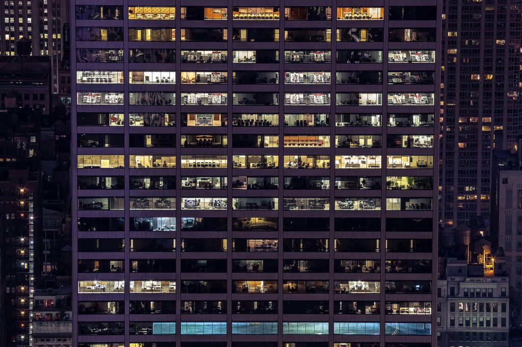 A side view of office blocks in Manhattan, New York, United States. It's mid-evening, and there's a purple haze. You can see rectangular office rooms lit up amongst other dotted dark rooms.