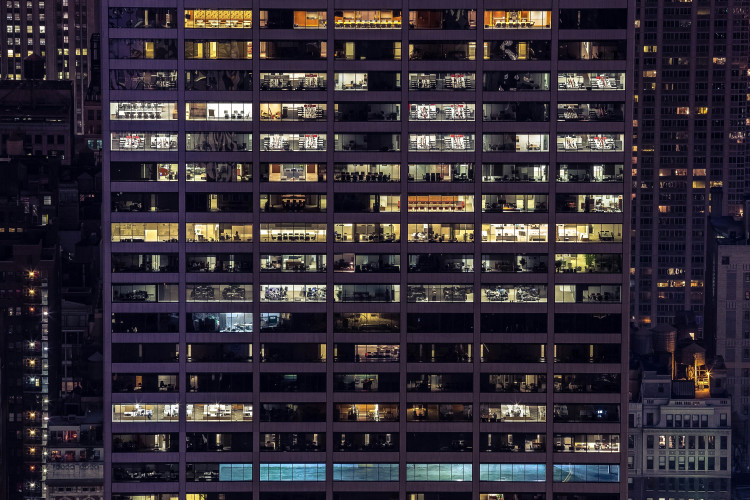 A side view of office blocks in Manhattan, New York, United States. It’s mid-evening, and there’s a purple haze. You can see rectangular office rooms lit up amongst other dotted dark rooms.