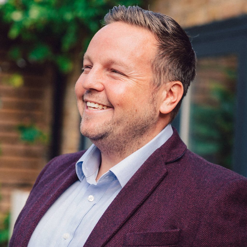 A side profile of Chris Morris slightly smiling. Chris has a light blue shirt on, and a maroon jacket.