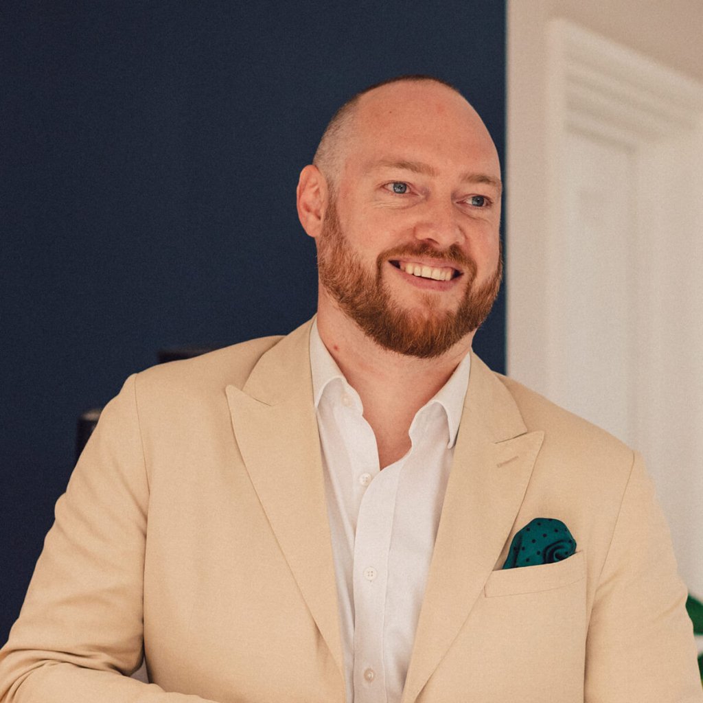Dan is smiling, looking away from the camera. He's a tall, broad-shouldered man with a shaved head and a thick ginger beard. He's wearing a cream jacket and dark green polka-dotted handkerchief in the top pocket.
