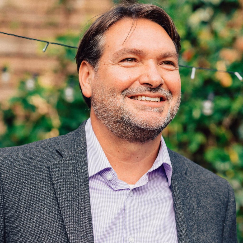 A side profile of Francisco smiling while he's looking slightly upwards. Francisco is wearing a mid blue checkered shirt and a dark-grey jacket. There's a blurred out brick wall covered in ivy behind him.
