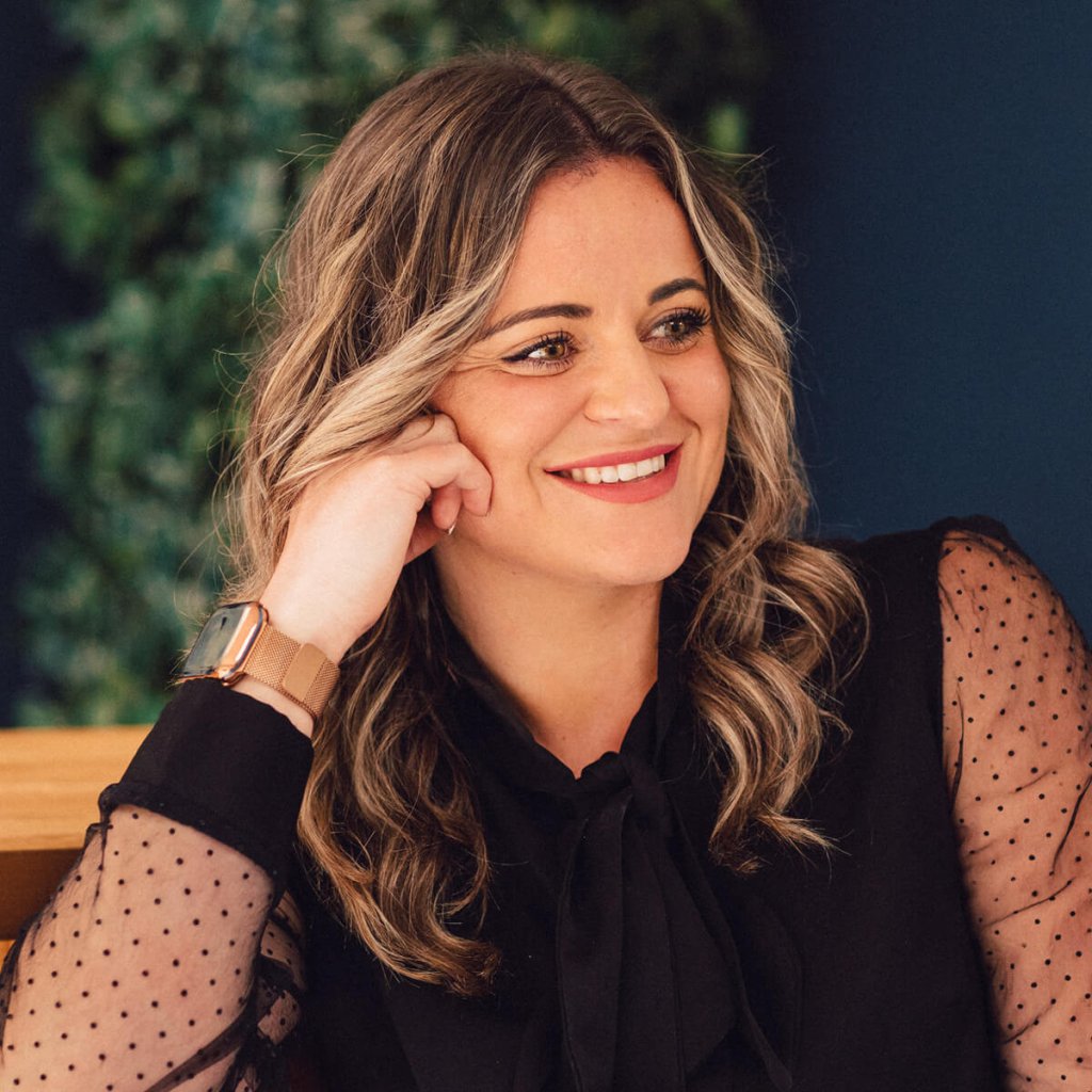 A side profile of Jo. She's smiling with a muted pink lipstick, blonde highlights and a black blouse with polka-dotted arms.