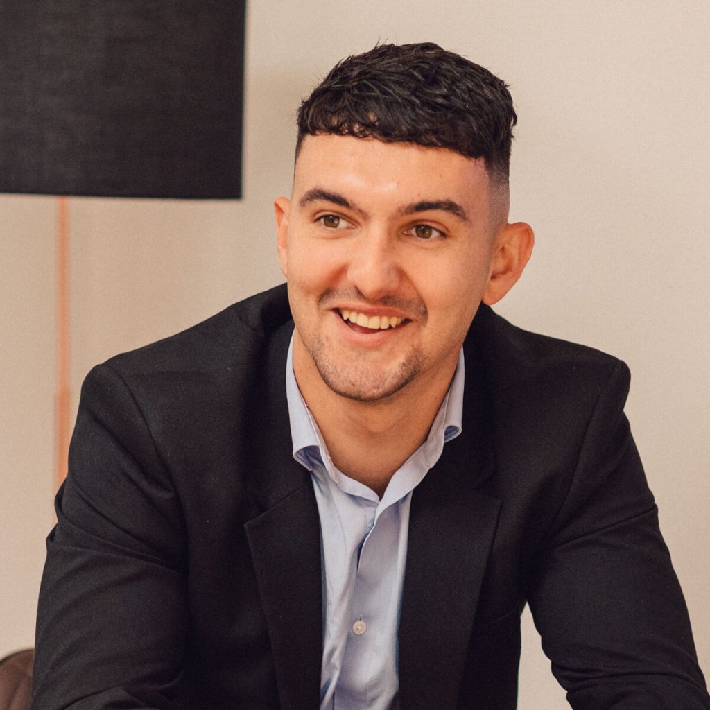 Jack's smiling cheekily, looking slightly off-camera. He's wearing a dark blazer jacket and light blue shirt.