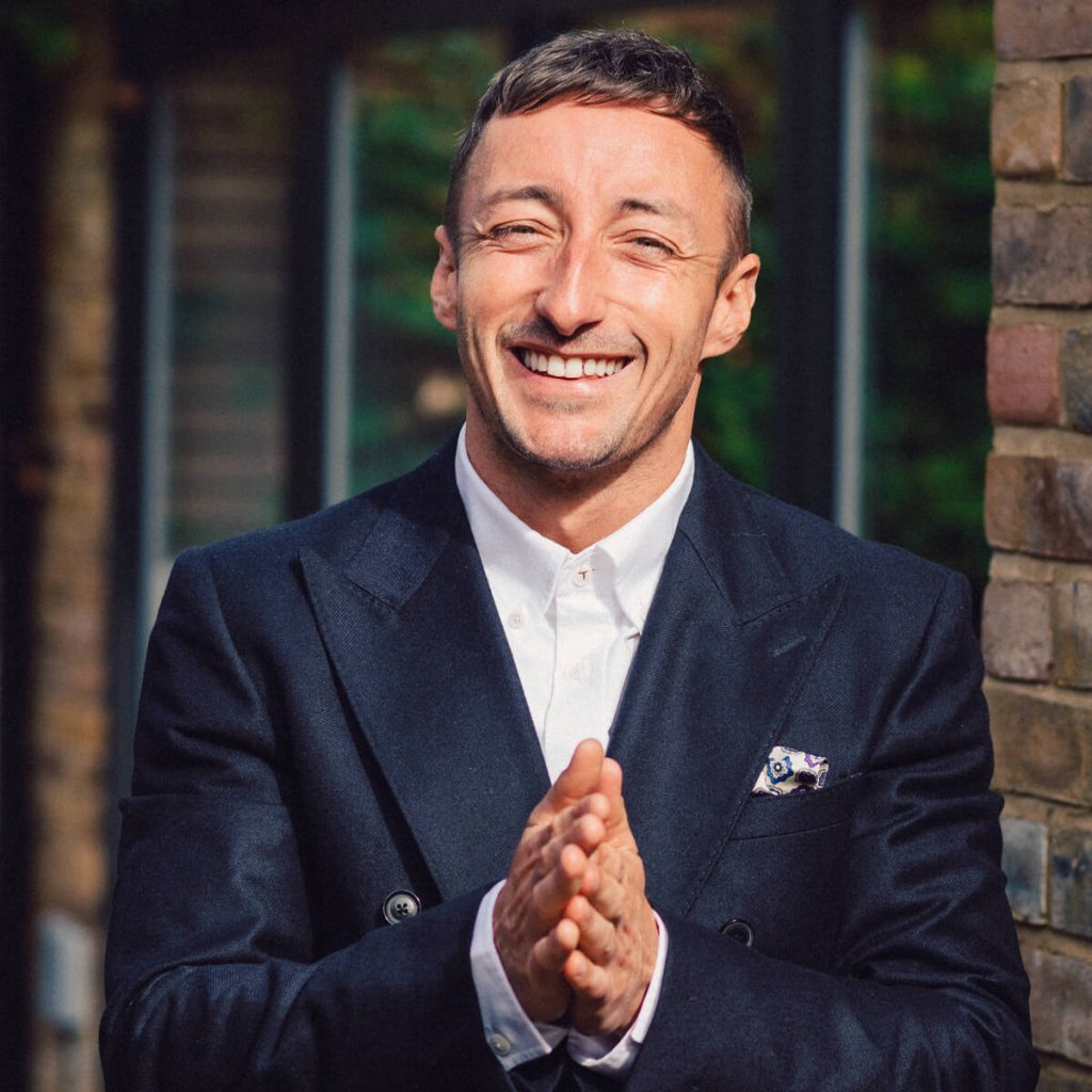 James McGregor smiling with his hands clasped together. James is wearing a smart blue tailored suit with a white shirt.