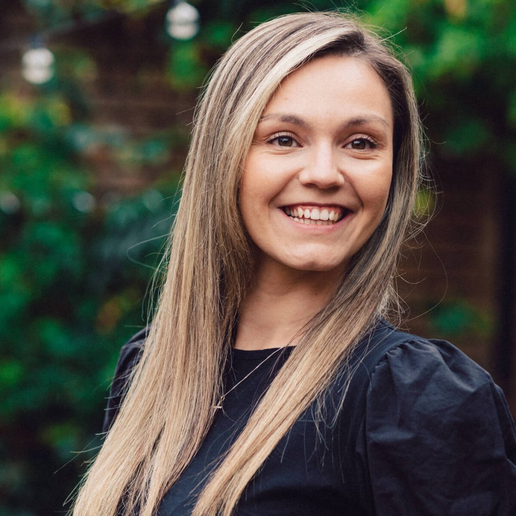 Lois is smiling looking slightly off-camera. She has a dark short-sleeved top on with a silver necklace and cross. Lois has brown eyes and blonde-streaked hair.